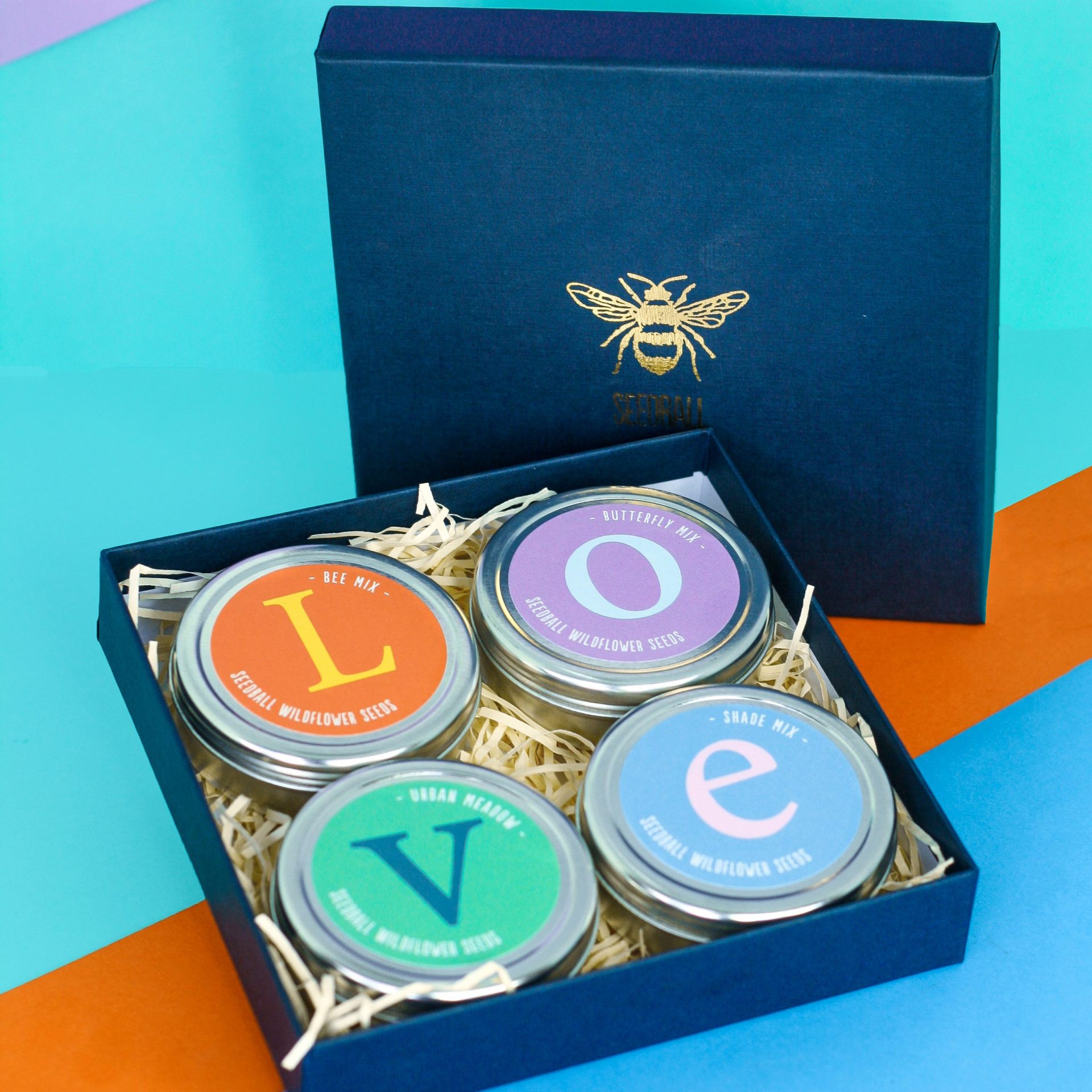 Image showing an open navy blue gift box with shredded paper packaging in and a gold bee on the lid. Within the shredded paper lies four round silver tins; each tin has a different coloured label. One is orange, one is purple, one is turquoise green and one is sky blue. The orange tin contains a yellow 'L' on the front, the purple tin a light blue 'O', the turquoise green tin a dark blue 'V', and the skyblue tin a purple 'E'. They are arranged in a square within the box and spells the word 'love'.